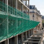 Filets de sécurité verts sur un bâtiment en construction à Paris, avec des ouvriers et des bâtiments haussmanniens en arrière-plan.