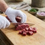Un chef ganté coupe de la viande de bœuf crue en dés sur une planche en bois, entouré d'ingrédients pour tartare.