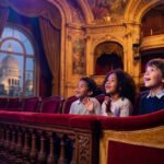 Trois enfants émerveillés dans une loge de théâtre parisien, le Sacré-Cœur visible par la fenêtre.